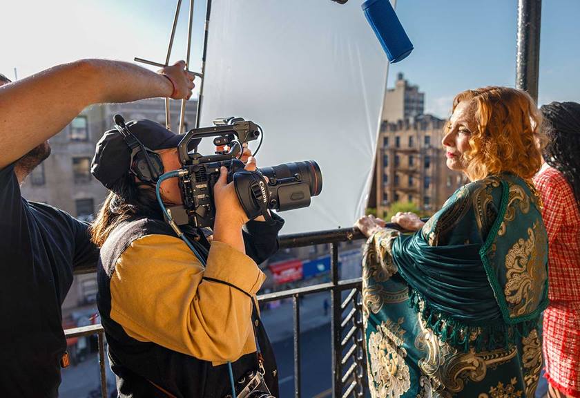 A woman resting camera on her shoulder, filming another woman on a balcony.