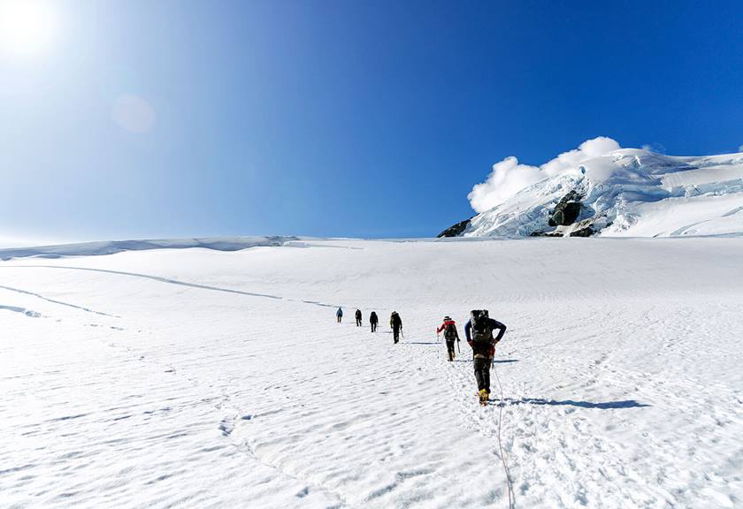 Trail of people traversing along icy plains with sun glare and blue skies