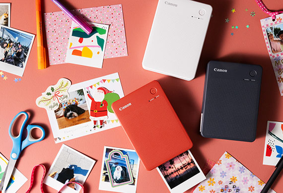 Flatlay image of three Canon Selphy Printers in red, white and black colours together with printed pictures and crafts